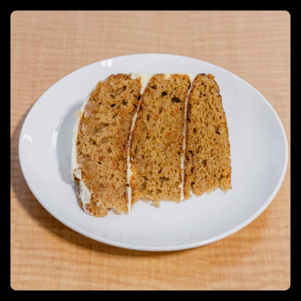 A slice of carrot cake with white frosting on a white plate, on a wooden table.