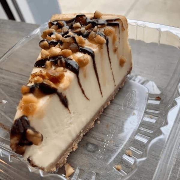 A slice of cheesecake with chocolate and caramel drizzle, topped with chopped nuts, in a plastic container.