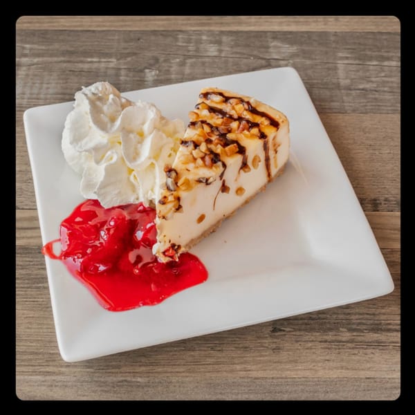Cheesecake slice with chocolate drizzle, nuts, whipped cream, and strawberry topping on a white plate.