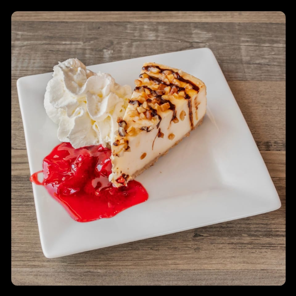 Cheesecake slice with chocolate drizzle, nuts, whipped cream, and strawberry topping on a white plate.