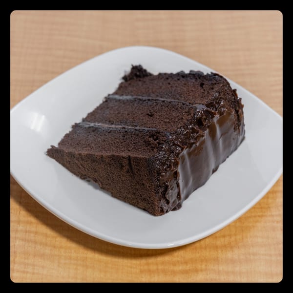 A slice of rich chocolate cake with dark frosting on a white square plate.
