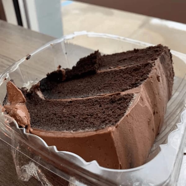 A large slice of rich chocolate cake with chocolate frosting in a clear plastic container.