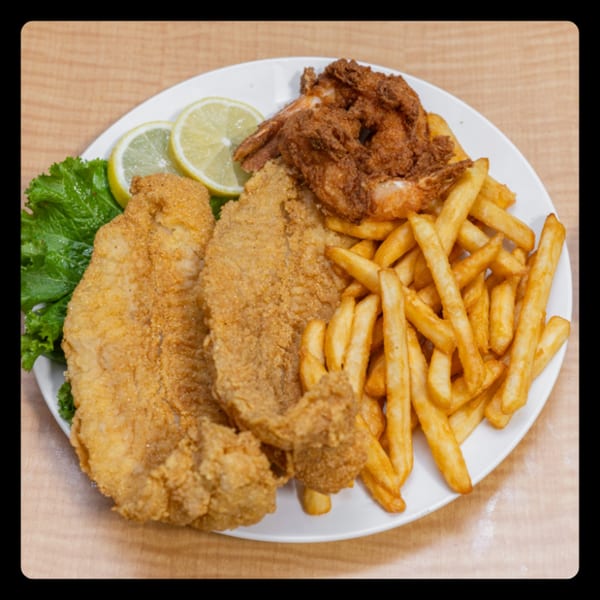 A white plate holds fried fish fillets, crispy shrimp, golden french fries, lettuce, and lemon slices.